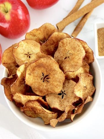 Air Fryer Apple Chips in white bowl with apples and cinnamon sticks in background